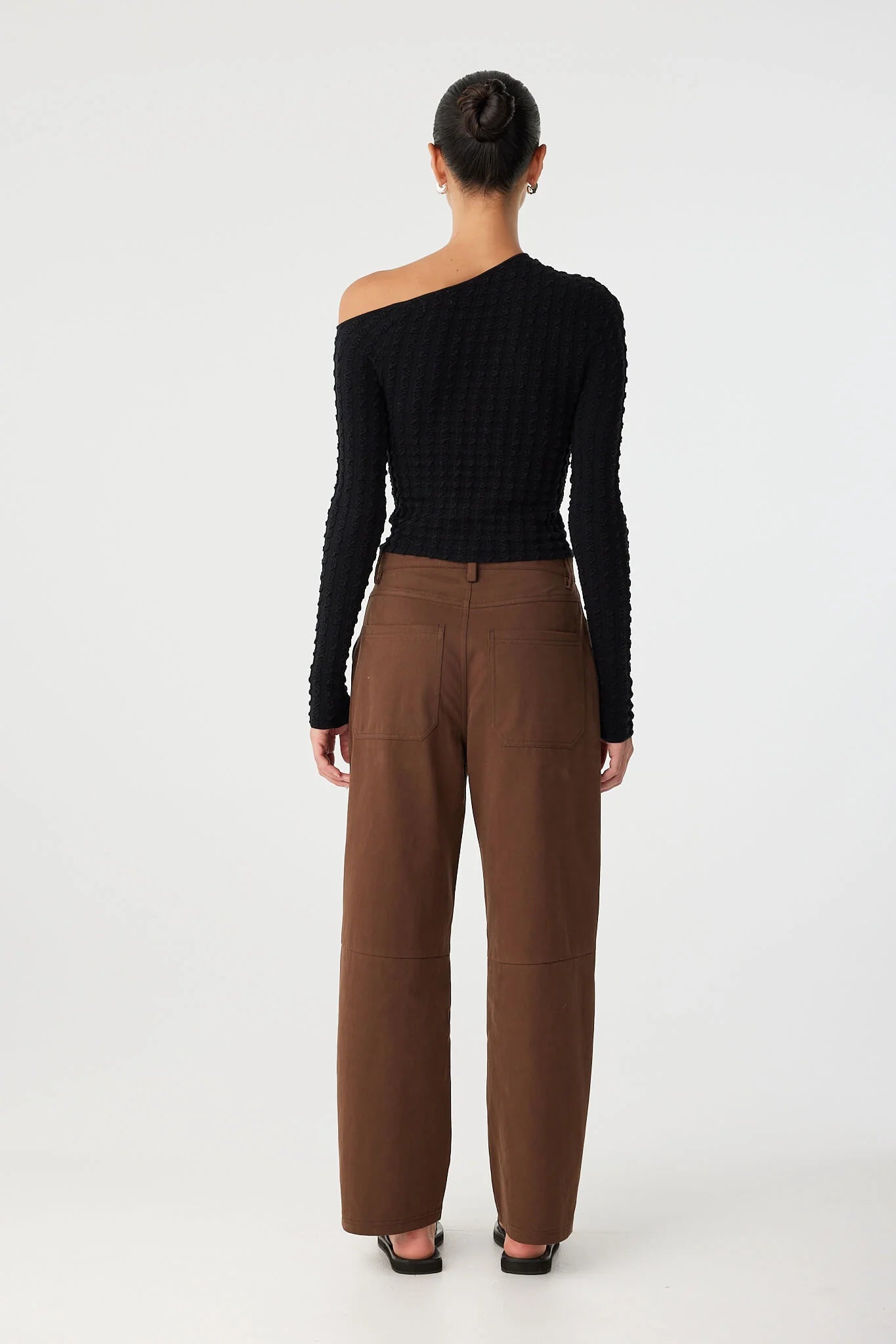 Person wearing a black one-shoulder top and brown pants on a white background