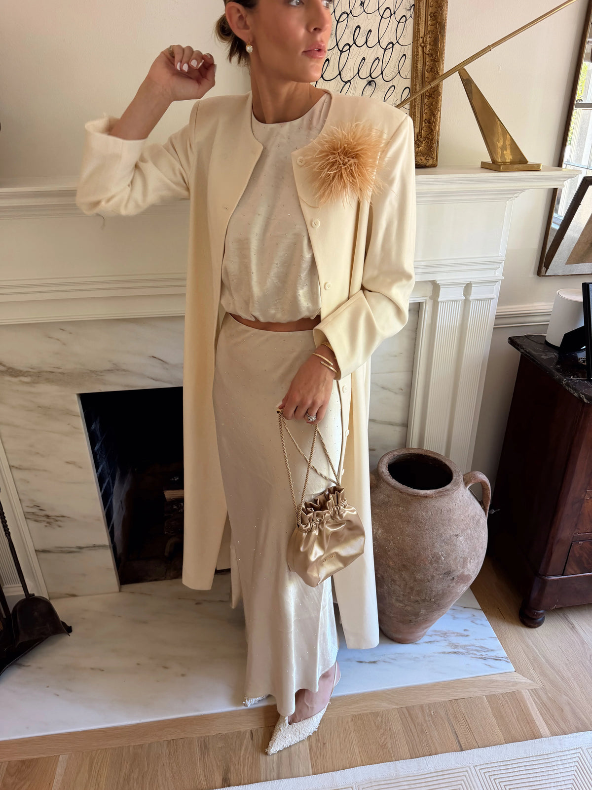 A woman models a cream-colored three-piece outfit consisting of a long-sleeve top, a matching midi skirt, and a long open-front cardigan. She accessorizes with a large beige feather embellishment on the cardigan, a small metallic gold drawstring bag, and white pointed-toe flats, standing in front of a white fireplace.