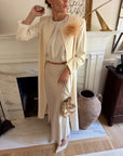 A woman models a cream-colored three-piece outfit consisting of a long-sleeve top, a matching midi skirt, and a long open-front cardigan. She accessorizes with a large beige feather embellishment on the cardigan, a small metallic gold drawstring bag, and white pointed-toe flats, standing in front of a white fireplace.