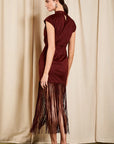 A woman stands in profile wearing a sleeveless high-neck dress in deep burgundy with a long fringe skirt, posed against a draped beige curtain backdrop.