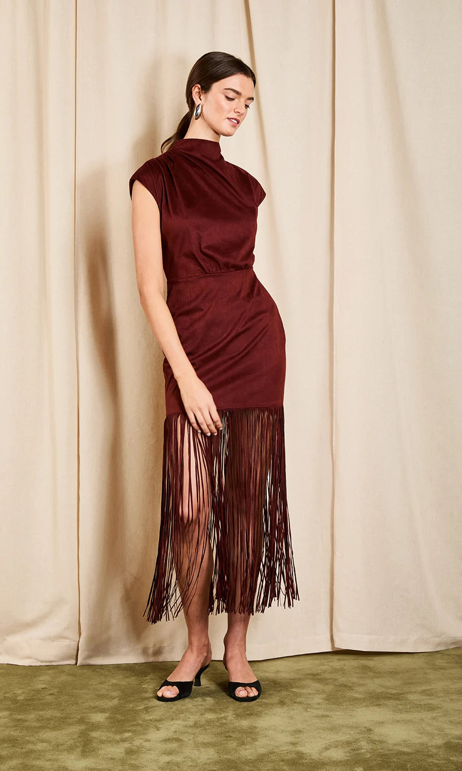 A woman models a sleeveless, high-neck burgundy dress with a draped neckline and a long fringe hem, standing against a beige curtain backdrop.