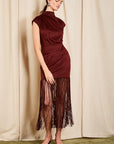 A woman models a sleeveless, high-neck burgundy dress with a draped neckline and a long fringe hem, standing against a beige curtain backdrop.