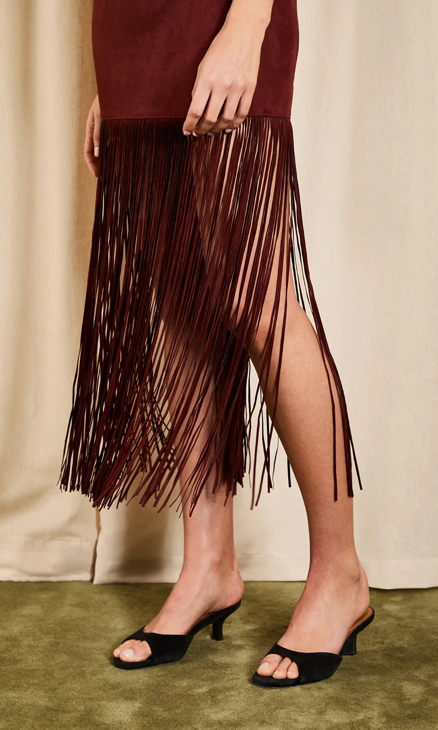A person wearing a dark burgundy fringe midi skirt with a side slit and black open-toe mule heels, standing against a neutral beige curtain backdrop.