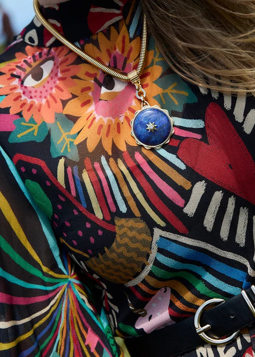 A close-up lifestyle shot of a person wearing a colorful, patterned blouse with a black belt and a gold necklace featuring a blue circular pendant.
