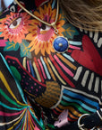 A close-up lifestyle shot of a person wearing a colorful, patterned blouse with a black belt and a gold necklace featuring a blue circular pendant.