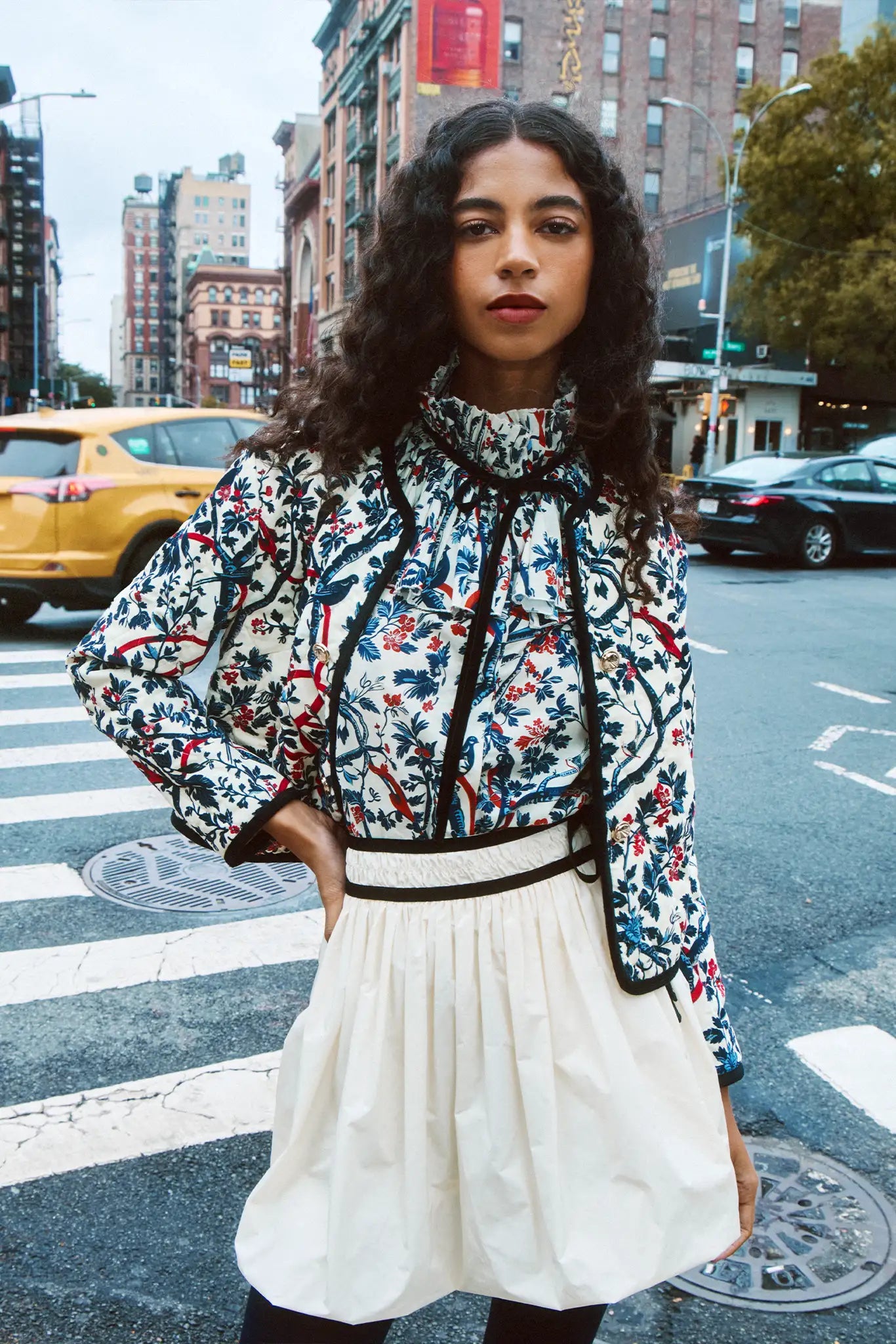 Woman in a floral blouse and white skirt standing on a city street.