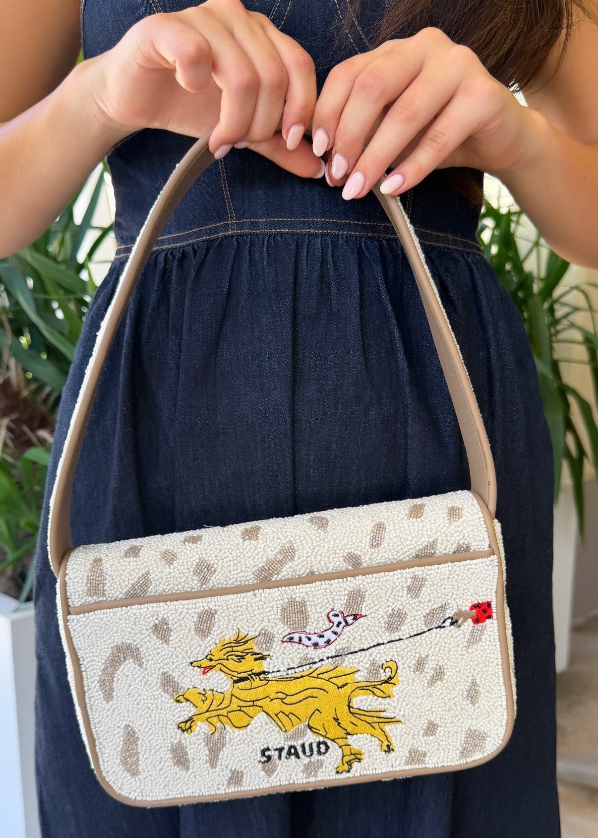 A woman in a dark denim dress holds a Staud shoulder bag with a cream and beige leopard print, featuring a yellow embroidered dog and the Staud logo.