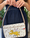A woman in a dark denim dress holds a Staud shoulder bag with a cream and beige leopard print, featuring a yellow embroidered dog and the Staud logo.