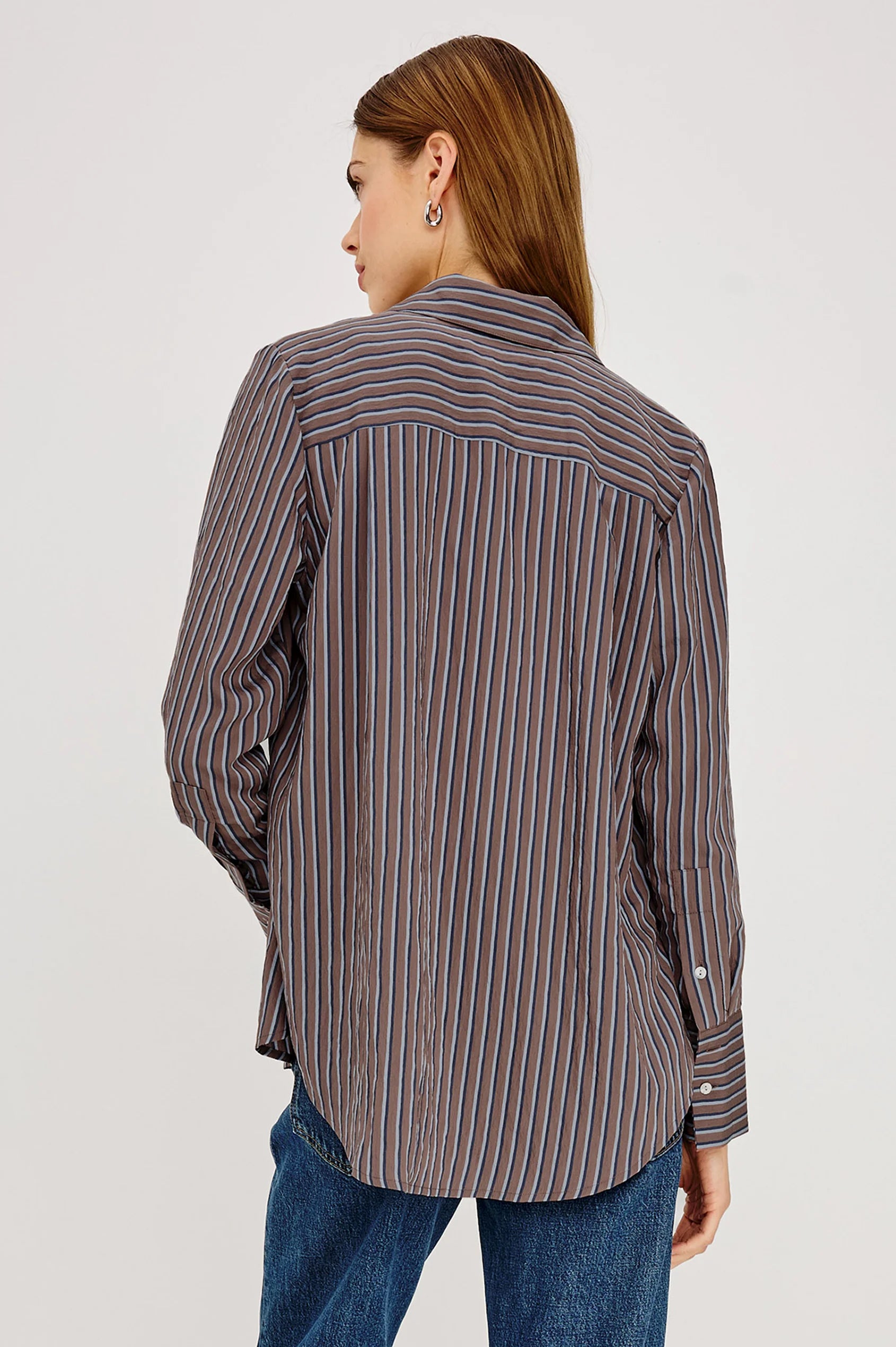 A woman shown from the back wearing a long-sleeve button-up shirt with vertical blue, brown, and white stripes, paired with blue jeans, against a plain light gray studio background.
