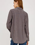 A woman shown from the back wearing a long-sleeve button-up shirt with vertical blue, brown, and white stripes, paired with blue jeans, against a plain light gray studio background.
