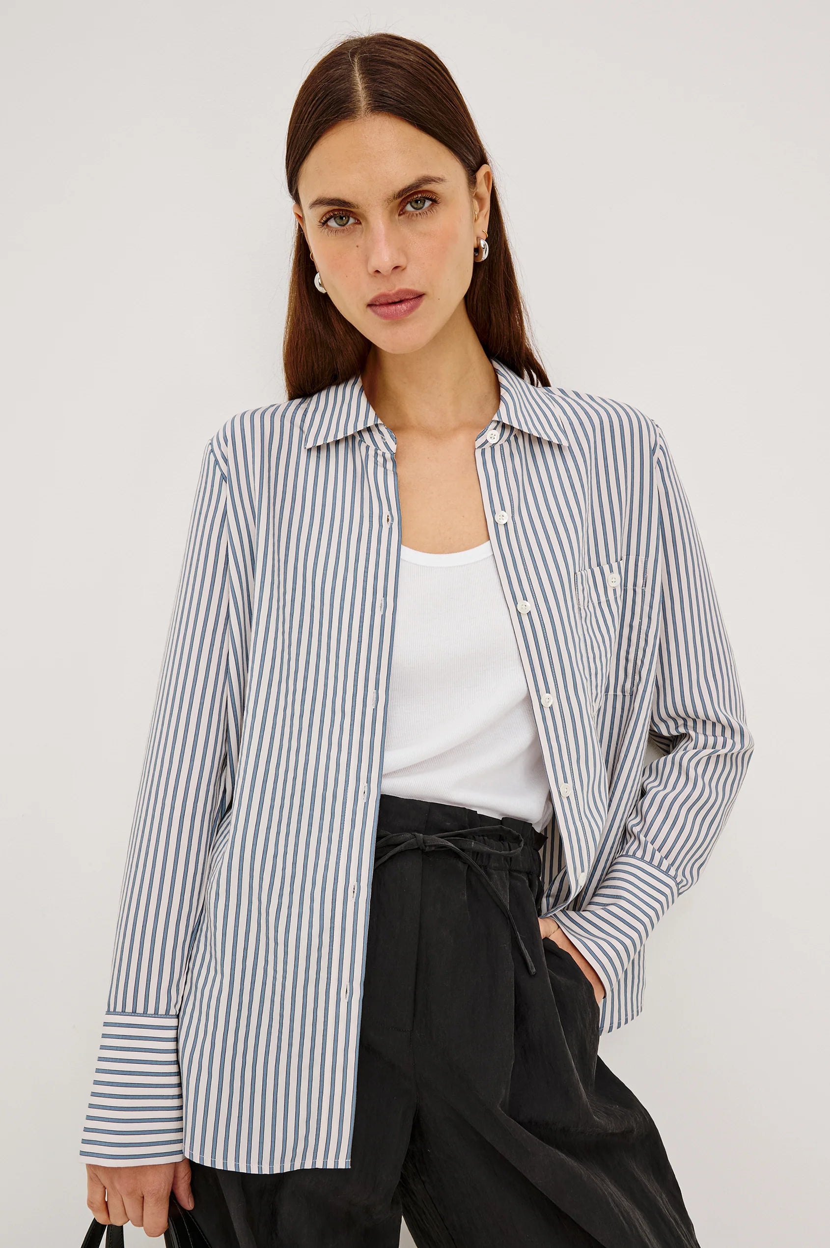 A woman modeling a blue and white vertically striped button-up shirt over a white tank top and black pants, photographed against a plain light gray studio background.