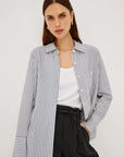 A woman modeling a blue and white vertically striped button-up shirt over a white tank top and black pants, photographed against a plain light gray studio background.
