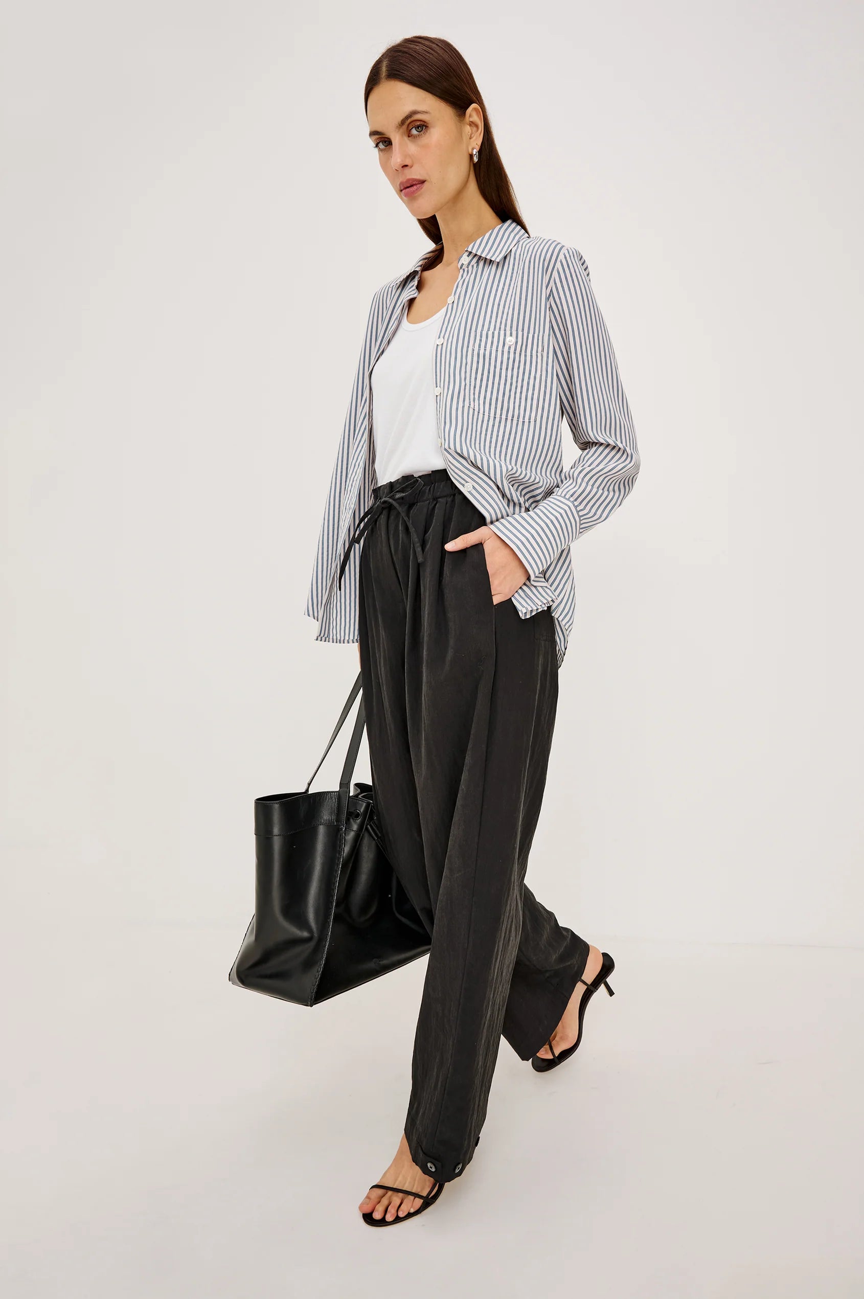 A woman models a stylish outfit consisting of a blue and white vertically striped button-down shirt, a white tank top, black wide-leg trousers, and black strappy heels, while carrying a black leather tote bag against a plain light gray studio background.
