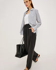A woman models a stylish outfit consisting of a blue and white vertically striped button-down shirt, a white tank top, black wide-leg trousers, and black strappy heels, while carrying a black leather tote bag against a plain light gray studio background.