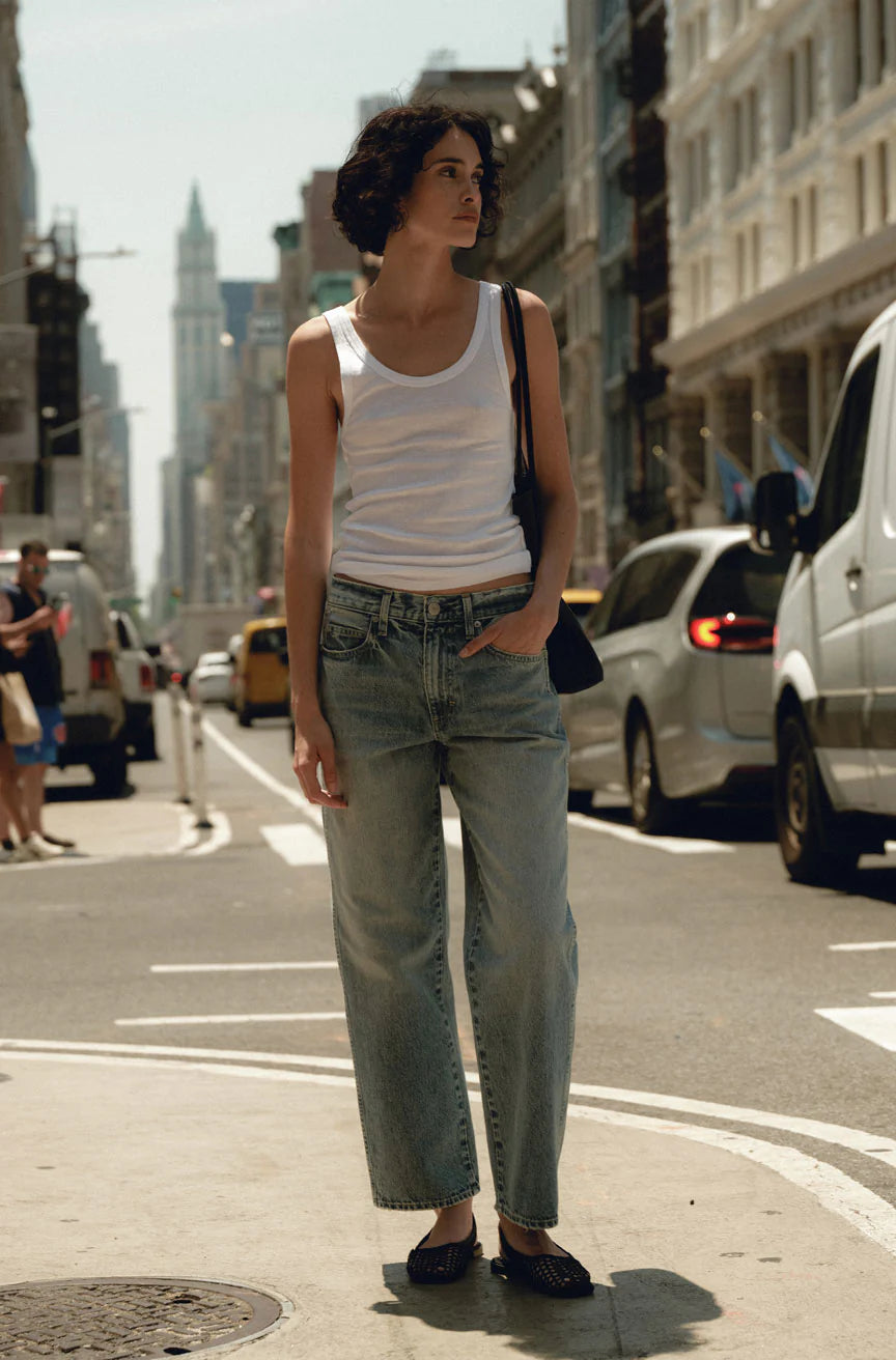 Woman standing on a city street wearing a white tank top and blue jeans.
