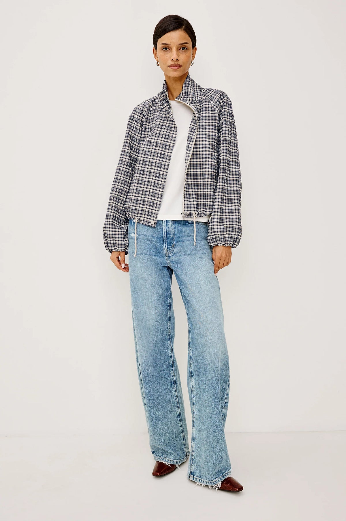 A model stands against a plain light background wearing a black and white plaid cropped jacket over a white top, light blue wide-leg jeans, and dark brown pointed shoes.