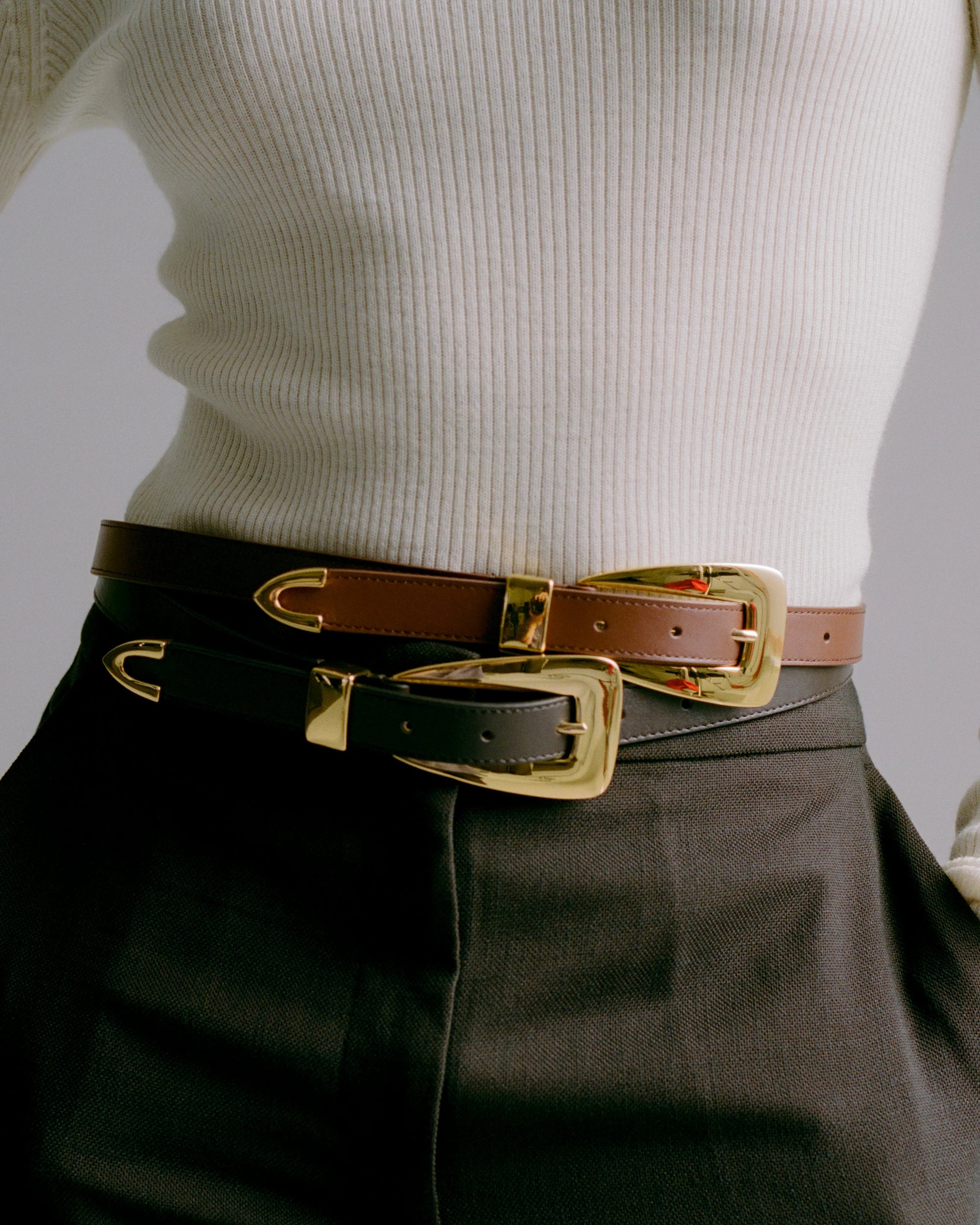 Person wearing a white ribbed sweater and dark pants with a brown belt and black belt featuring gold buckles.