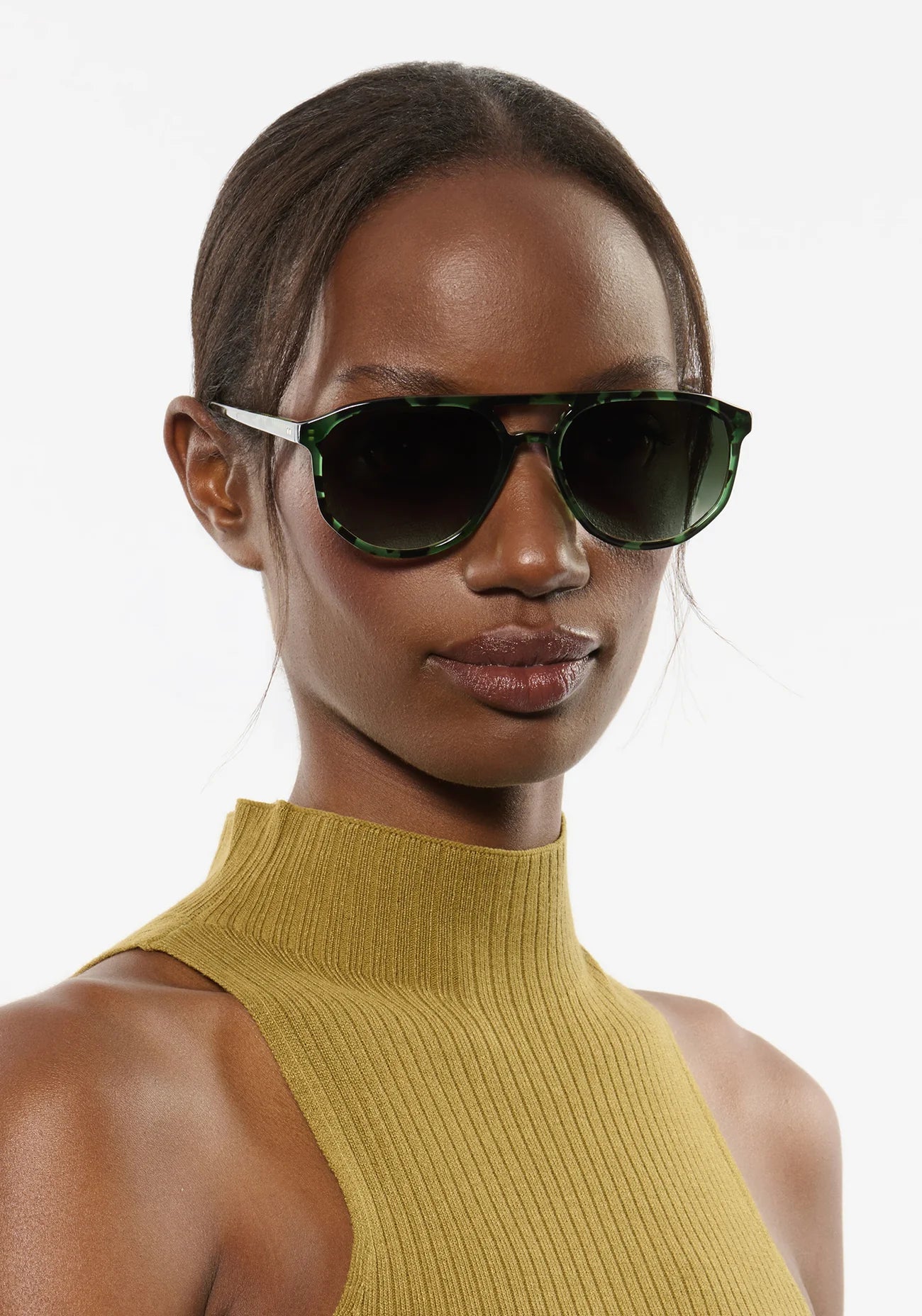 Woman wearing dark sunglasses and a mustard yellow top against a white background