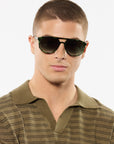 A man with a buzz cut wearing dark green aviator sunglasses with sunglasses and a textured olive green polo shirt, set against a plain white background