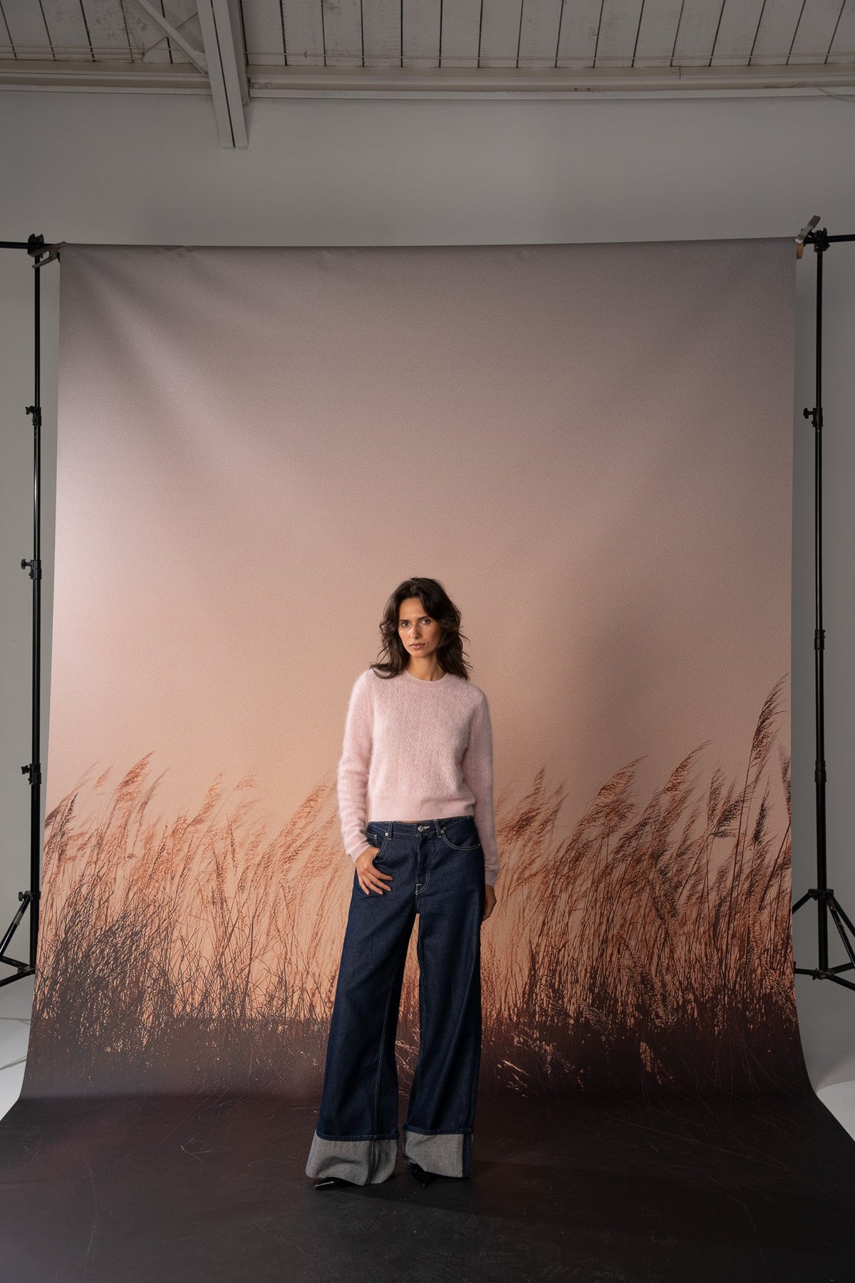 A woman stands in front of a large printed photography backdrop featuring a soft pink sky and brown reeds, wearing a light pink knit sweater and dark wide-leg jeans.
