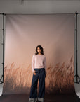 A woman stands in front of a large printed photography backdrop featuring a soft pink sky and brown reeds, wearing a light pink knit sweater and dark wide-leg jeans.