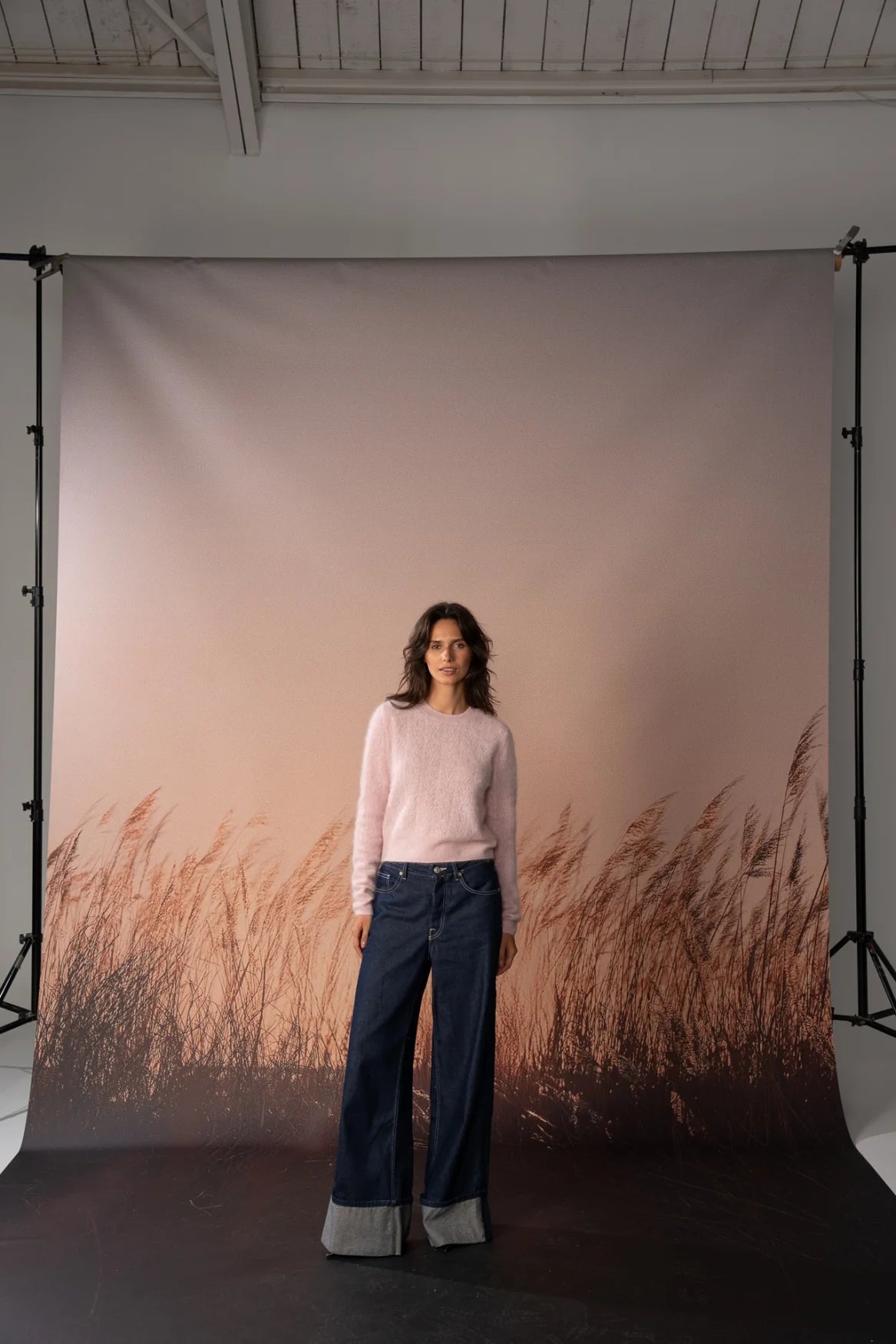 A woman stands in front of a large studio backdrop featuring a muted pink sky and a brown grassy field, wearing a light pink long-sleeve top and dark blue wide-leg jeans with gray cuffed hems.