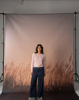 A woman stands in front of a large studio backdrop featuring a muted pink sky and a brown grassy field, wearing a light pink long-sleeve top and dark blue wide-leg jeans with gray cuffed hems.