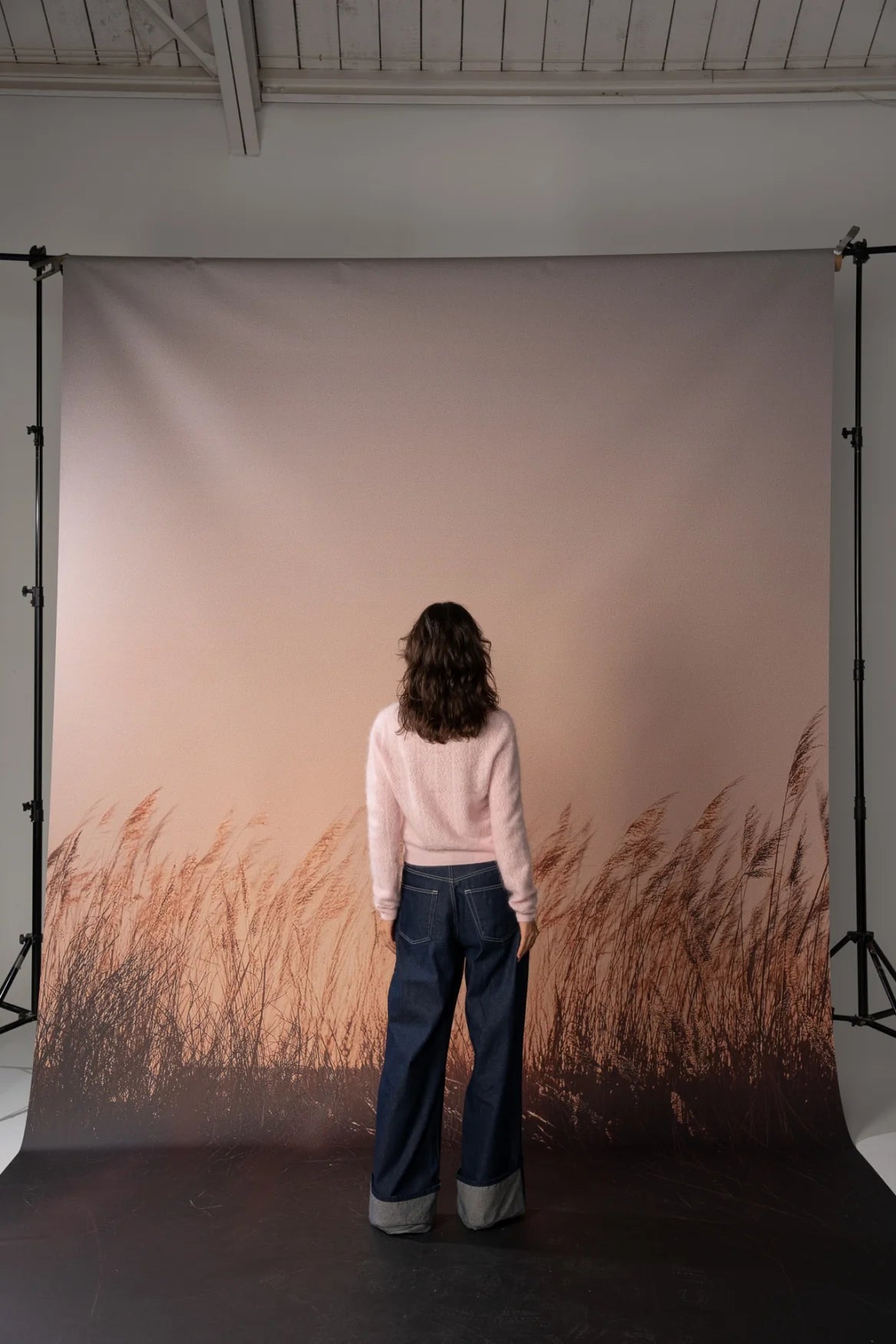 A person stands with their back to the camera in front of a large photography backdrop featuring a warm beige sky and a brown grassy field. The backdrop is mounted on a studio stand system.