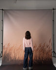 A person stands with their back to the camera in front of a large photography backdrop featuring a warm beige sky and a brown grassy field. The backdrop is mounted on a studio stand system.