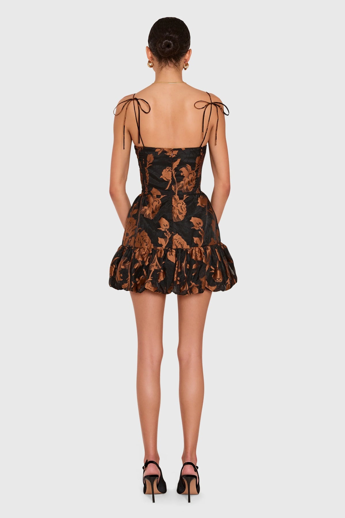 A model viewed from the back wearing a short black dress with a brown floral jacquard pattern, thin shoulder tie straps, and a ruffled hem, paired with black high heels against a light gray background.