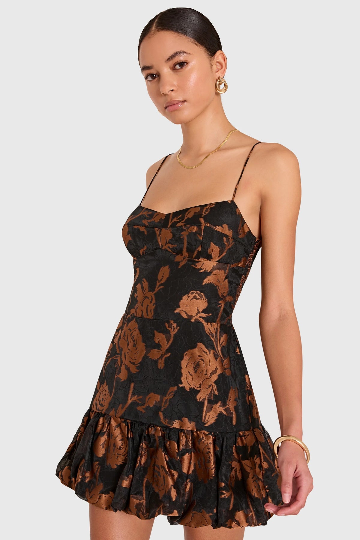 A woman modeling a black spaghetti-strap mini dress with a bronze-brown floral print, set against a solid light gray background.