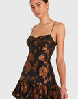 A woman modeling a black spaghetti-strap mini dress with a bronze-brown floral print, set against a solid light gray background.