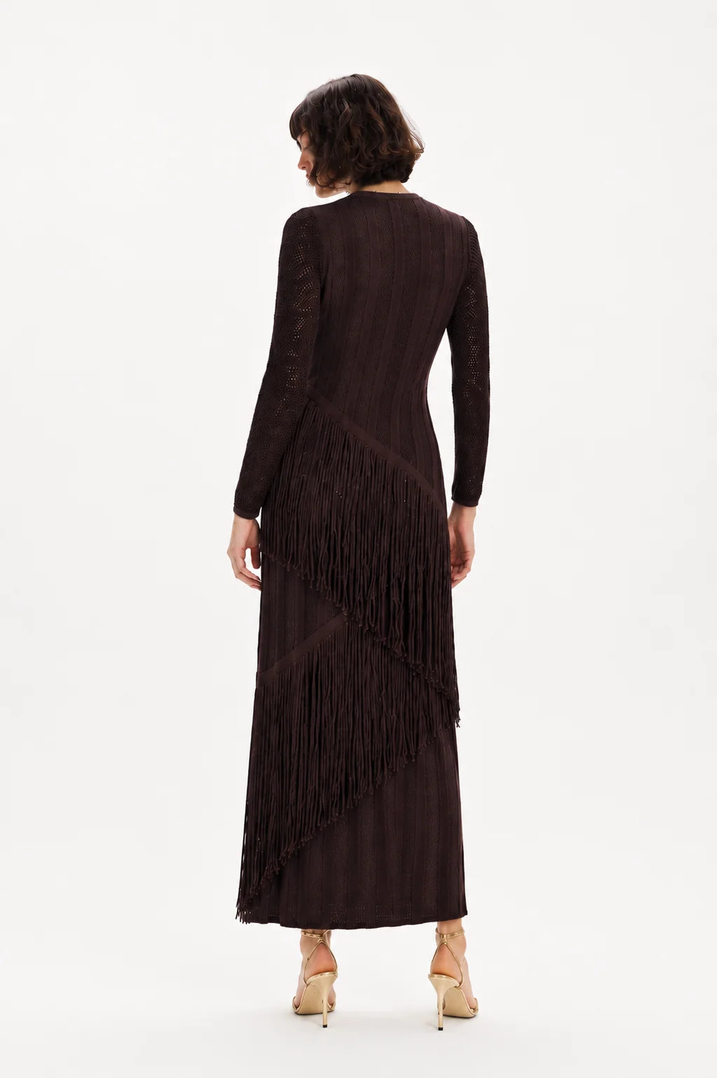 A woman viewed from the back wearing a long-sleeve dark brown fringe dress and beige high heels against a plain white studio background.