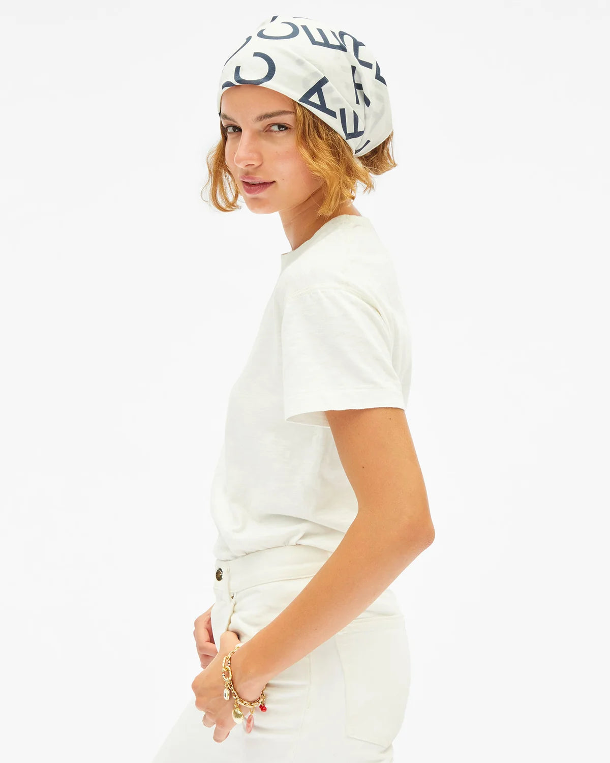 A woman wearing a white crewneck t-shirt, white pants, and a white headscarf with navy blue graphic lettering, photographed against a solid white background.