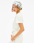 A woman wearing a white crewneck t-shirt, white pants, and a white headscarf with navy blue graphic lettering, photographed against a solid white background.
