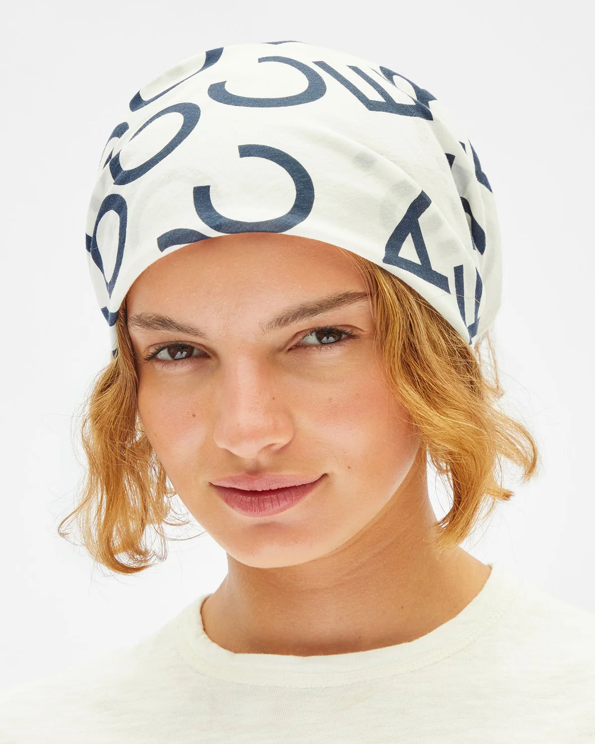 A close-up of a model wearing a white fabric headscarf with a navy blue abstract graphic pattern, set against a plain white background.