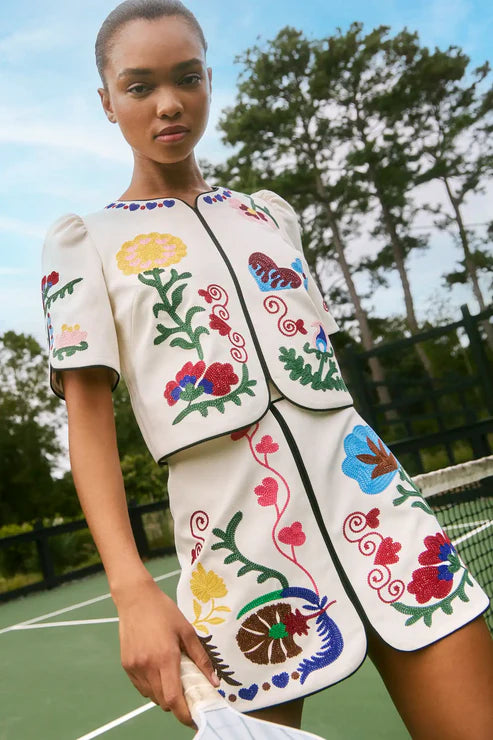 Woman wearing a white outfit with colorful floral embroidery on a tennis court.