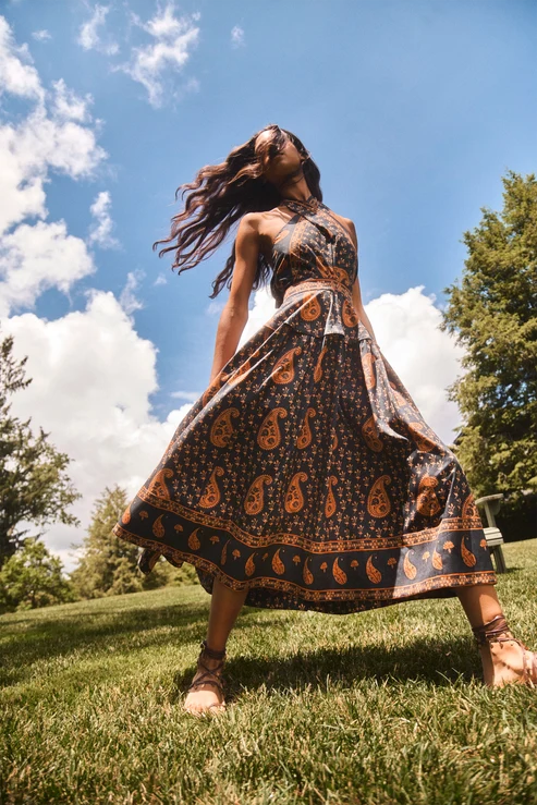 A woman wearing a flowing paisley maxi dress stands on a grassy lawn under a blue sky with clouds. The dress has a dark base with orange and beige paisley motifs and a matching patterned border.