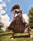 A woman wearing a flowing paisley maxi dress stands on a grassy lawn under a blue sky with clouds. The dress has a dark base with orange and beige paisley motifs and a matching patterned border.