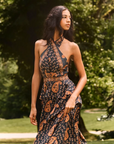 A woman wearing a sleeveless halter-neck maxi dress with a dark navy blue paisley and floral print, standing outdoors in a park with lush green trees in the background.
