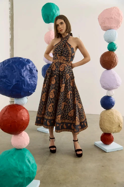 A woman stands in a studio wearing a dark halter-neck midi dress with a brown paisley pattern. The background features colorful sculptural paper ball columns.