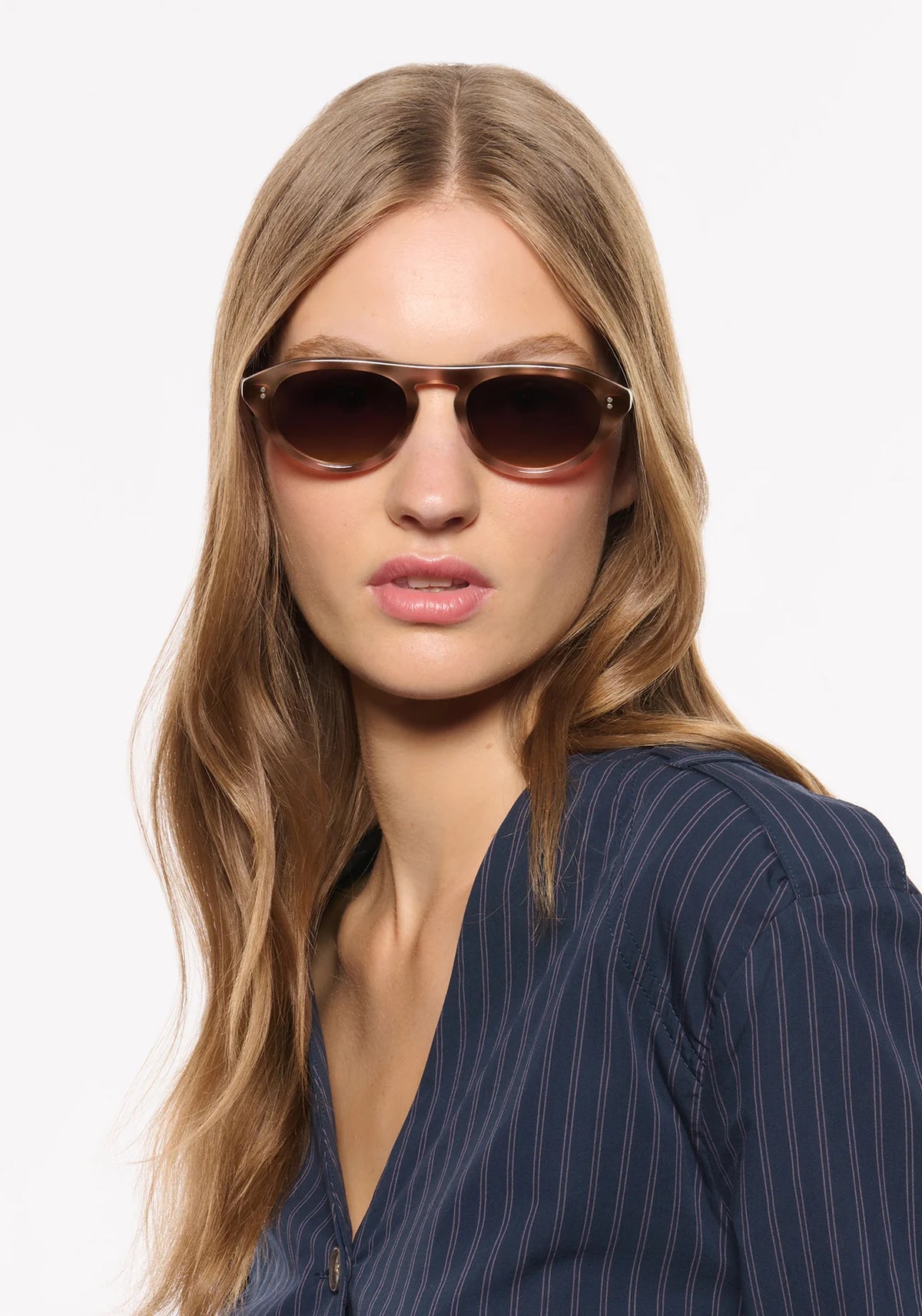 A woman with long blonde hair wearing dark brown cat-eye sunglasses and a navy blue pinstripe top, photographed against a clean white background.