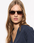 A woman with long blonde hair wearing dark brown cat-eye sunglasses and a navy blue pinstripe top, photographed against a clean white background.