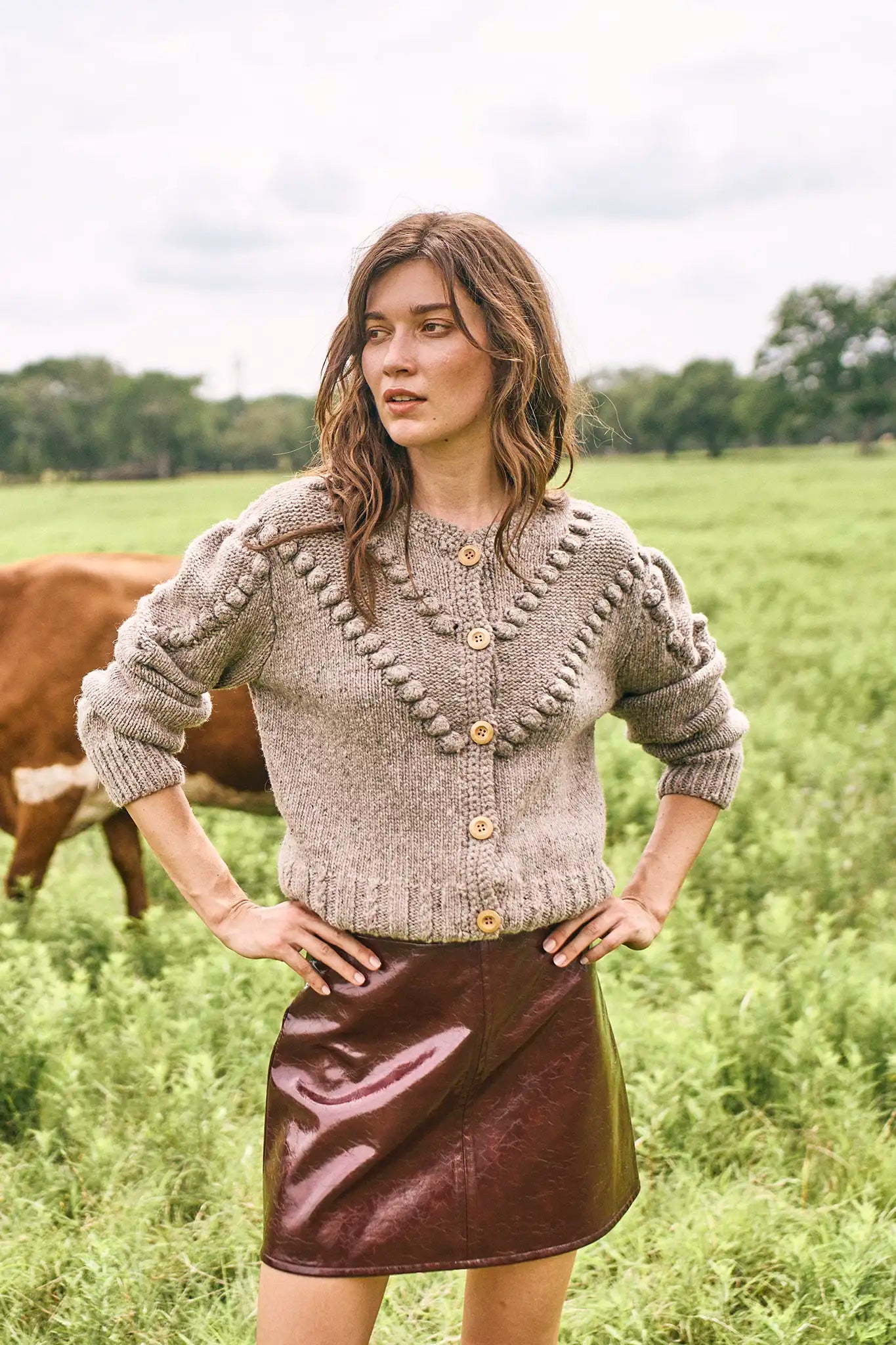 Woman wearing a textured sweater and leather skirt standing in a field with cows.