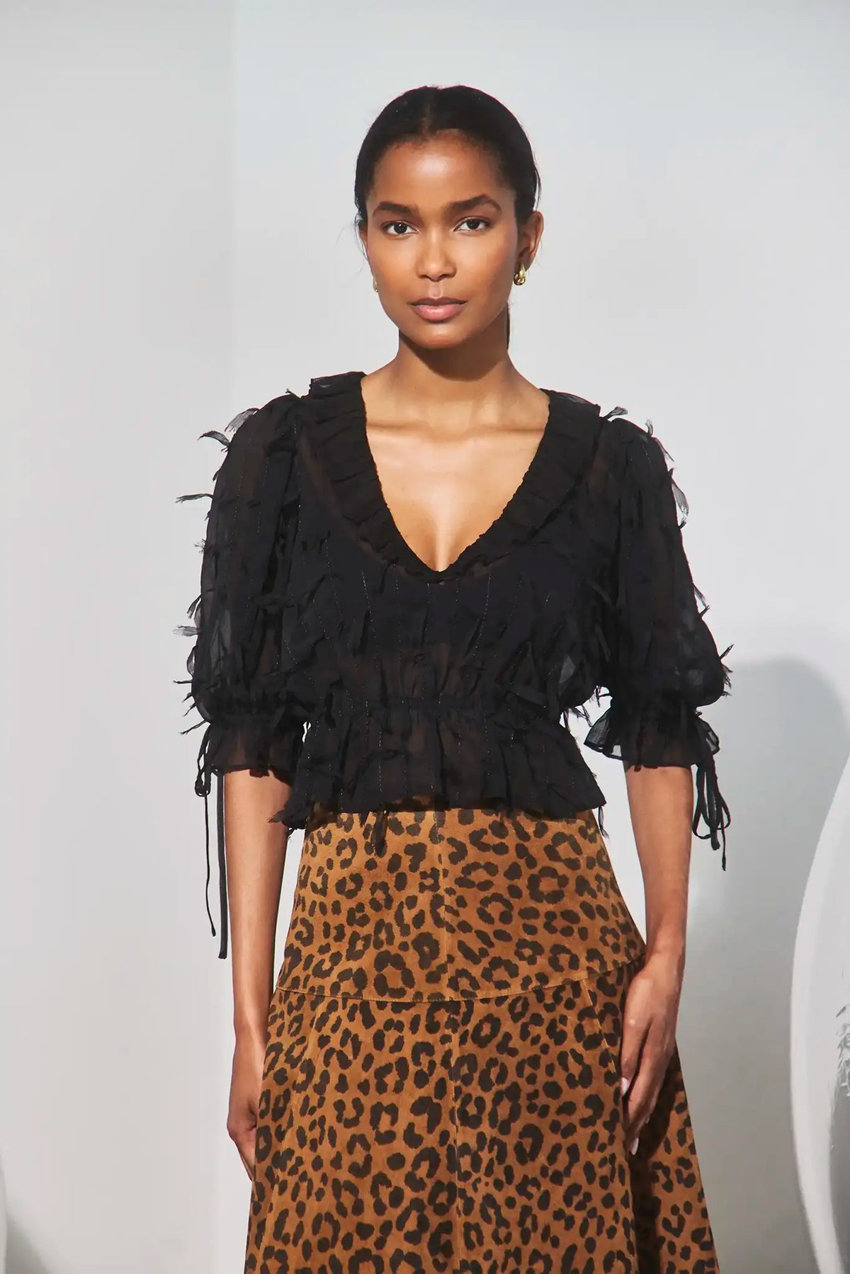 A model wearing a black ruffled short-sleeve blouse with tie details and a brown leopard-print skirt, standing against a light gray background.