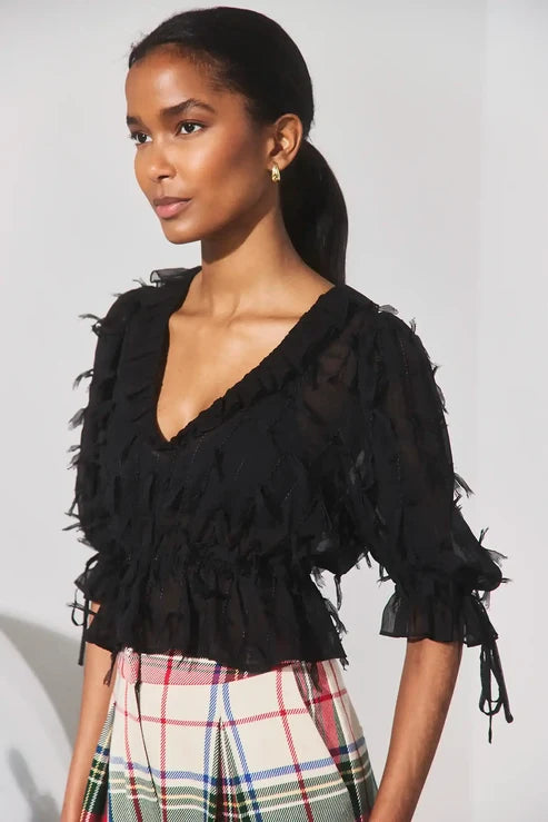 A woman wearing a black sheer ruffled blouse with tie details on the sleeves, paired with a plaid skirt, photographed against a plain light background.