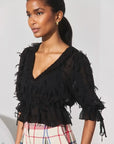 A woman wearing a black sheer ruffled blouse with tie details on the sleeves, paired with a plaid skirt, photographed against a plain light background.
