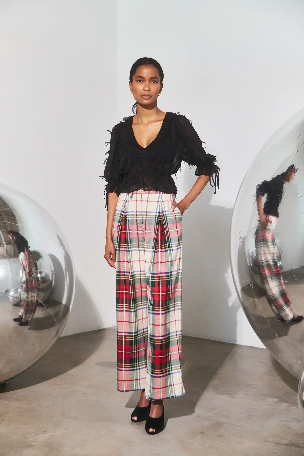 A model stands in a minimalist studio setting wearing a black fringed top and high-waisted plaid trousers in red, green, and white. She is framed by two large reflective silver spheres on either side of the background.
