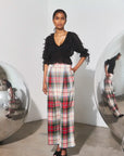 A model stands in a minimalist studio setting wearing a black fringed top and high-waisted plaid trousers in red, green, and white. She is framed by two large reflective silver spheres on either side of the background.