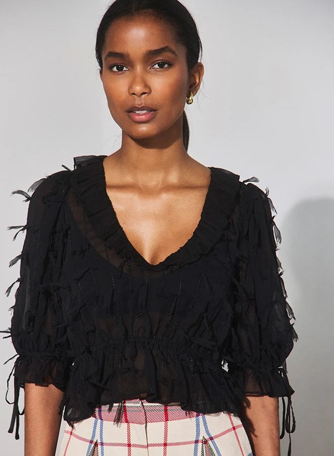 A model wearing a black sheer ruffled blouse with tiered puff sleeves and a deep V-neck, paired with a pink, white, and blue plaid skirt, against a plain light gray background.
